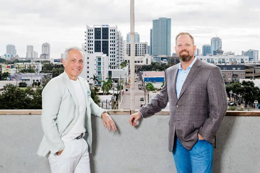 Two men standing on a rooftop and posing for a photo. the pair are wearing blazers