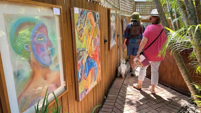 Two people walk down the brick pathway of an outdoor art exhibit, featuring colorful abstract paintings displayed on a wooden fence.