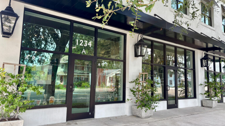 A modern gallery storefront with large glass windows and black-framed doors. The exterior features white walls, black awnings, wall-mounted lanterns, and potted plants along a clean sidewalk.