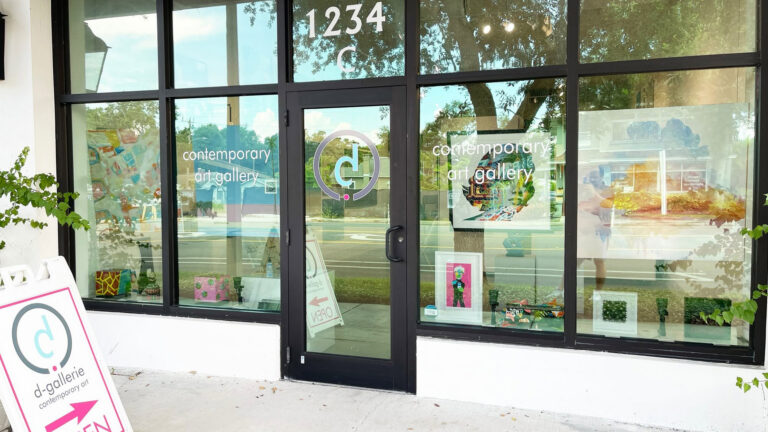 A contemporary art gallery storefront with large glass windows and a black door, displaying colorful artwork and signage with the gallery's logo and name.