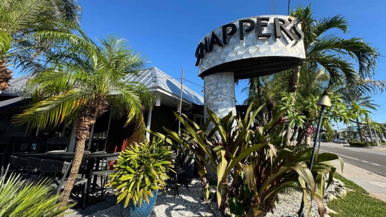 The outside seating area for Snappers Sea Grill, filled with tropical plants and palm trees under a clear blue sky.