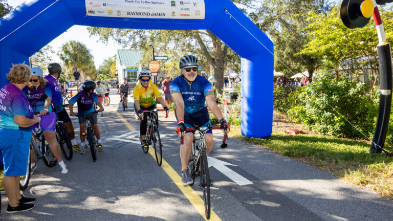 Cyclists participate in the 2025 Gulf Coast Bike Trek, passing under a blue inflatable arch with sponsor logos on a sunny day with spectators along the side.